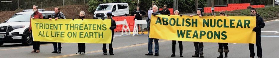 photo showing people block a road with banners that read "Trident threatens all life on earth" and "Abolish nuclear weapons."